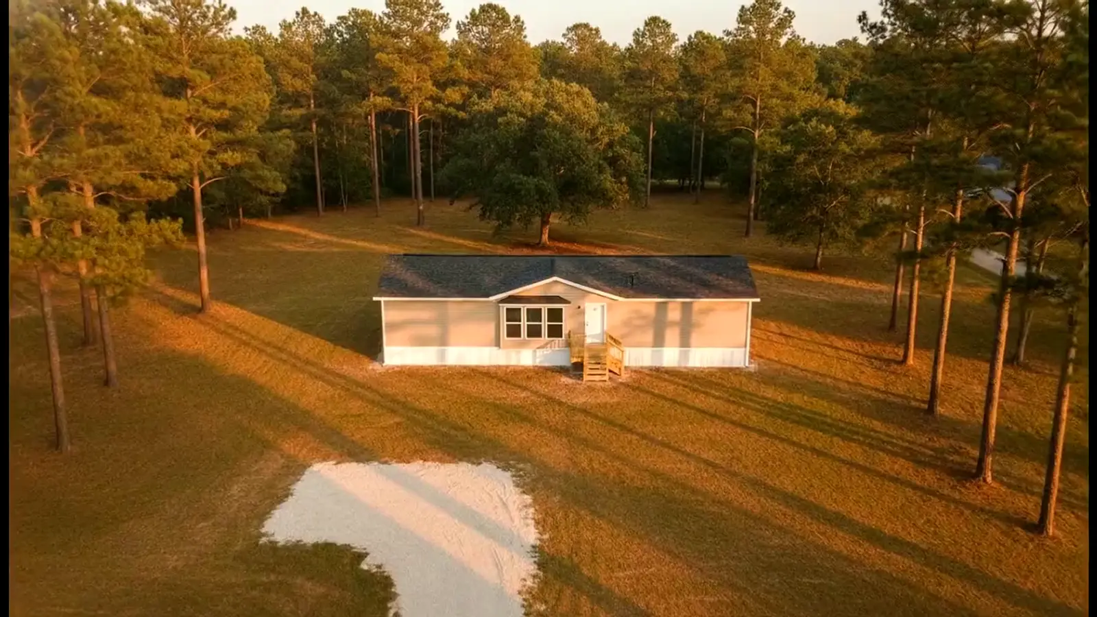 Vista aérea de una casa móvil de doble ancho instalada en su propio terreno entre los pinos del este de Texas al atardecer, con faldón instalado, escalones de madera y entrada de grava